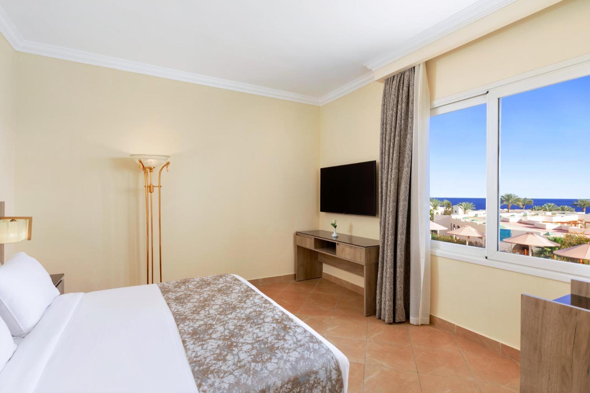 Pickalbatros Royal Grand Sharm - Adults Friendly 16 Years Plus Exteriör bild The photo shows a hotel room with a modern and minimalistic design. There is a comfortable bed with white linens and a decorative gray throw at the foot. A large window offers a view of the outdoors, possibly with a glimpse of the sea or beach. A fla