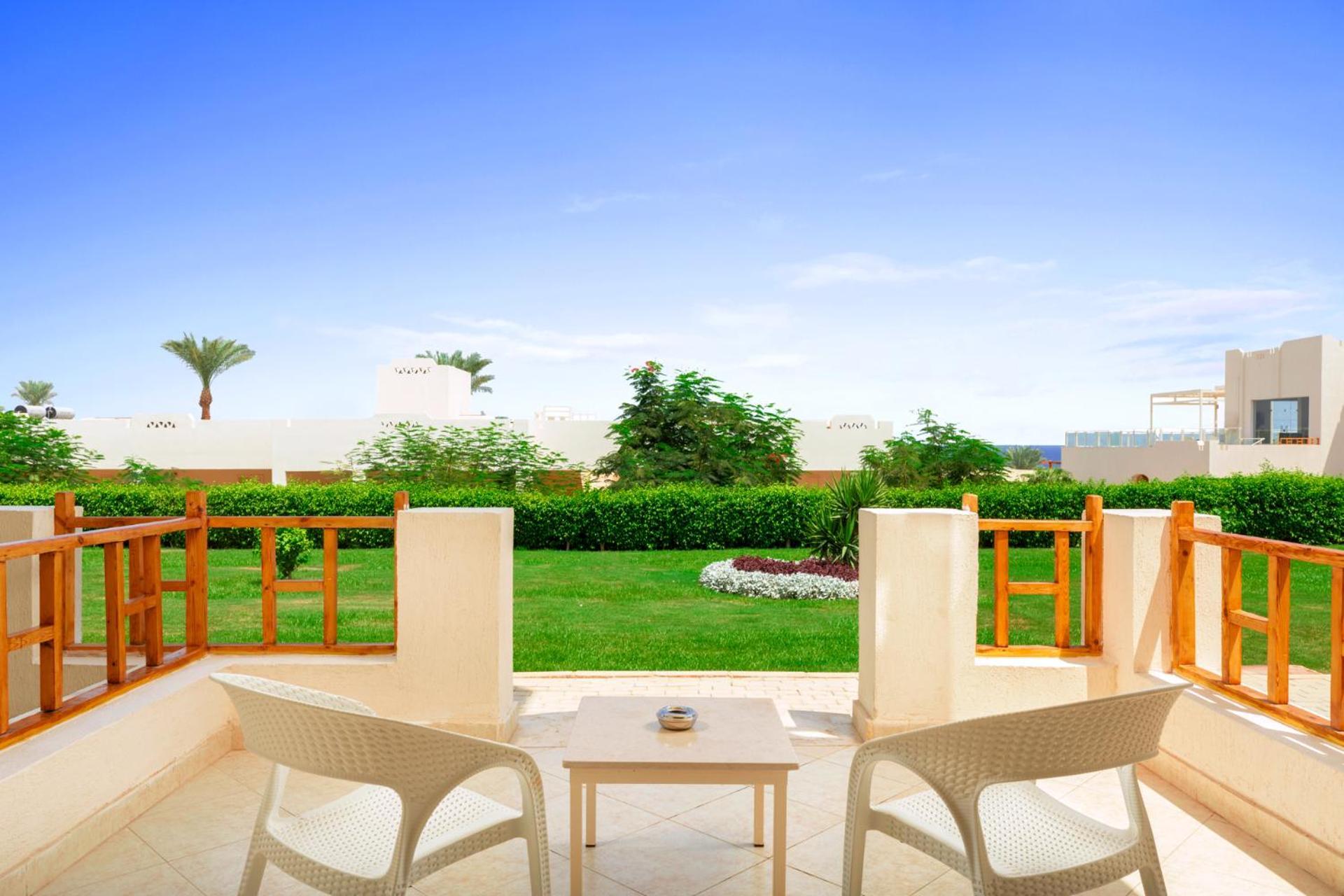 Pickalbatros Royal Grand Sharm - Adults Friendly 16 Years Plus Exteriör bild The photo depicts a serene outdoor balcony or patio area. It features two white chairs and a small table, set against a backdrop of lush green grass and garden plants. In the distance, there are some palm trees and clear blue skies, suggesting a warm