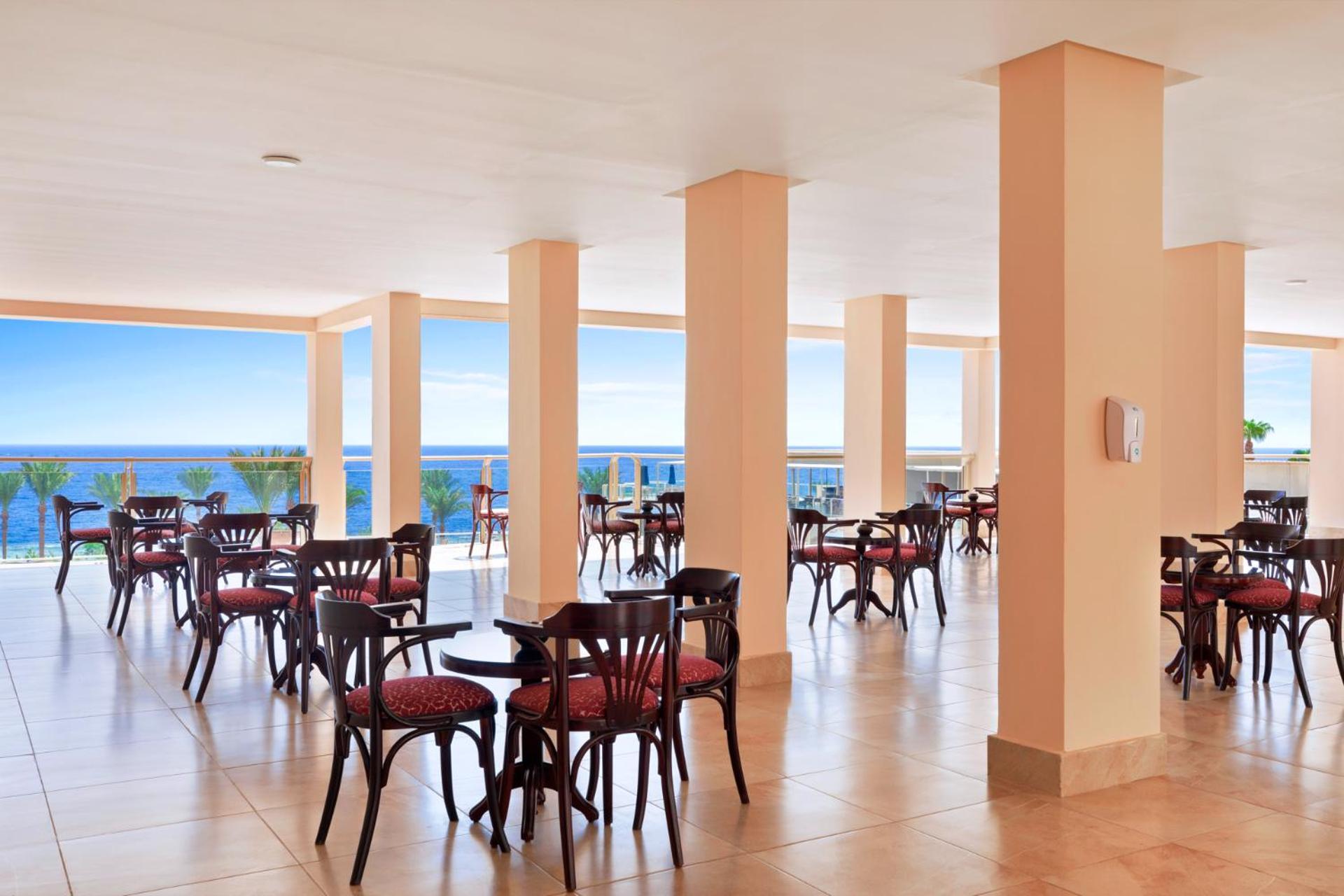 Pickalbatros Royal Grand Sharm - Adults Friendly 16 Years Plus Exteriör bild The image shows a dining area or café with several round wooden tables and chairs, featuring red upholstery. The space is well-lit, with large windows that allow a view of the ocean in the background, contributing to a bright and open atmosphere. The