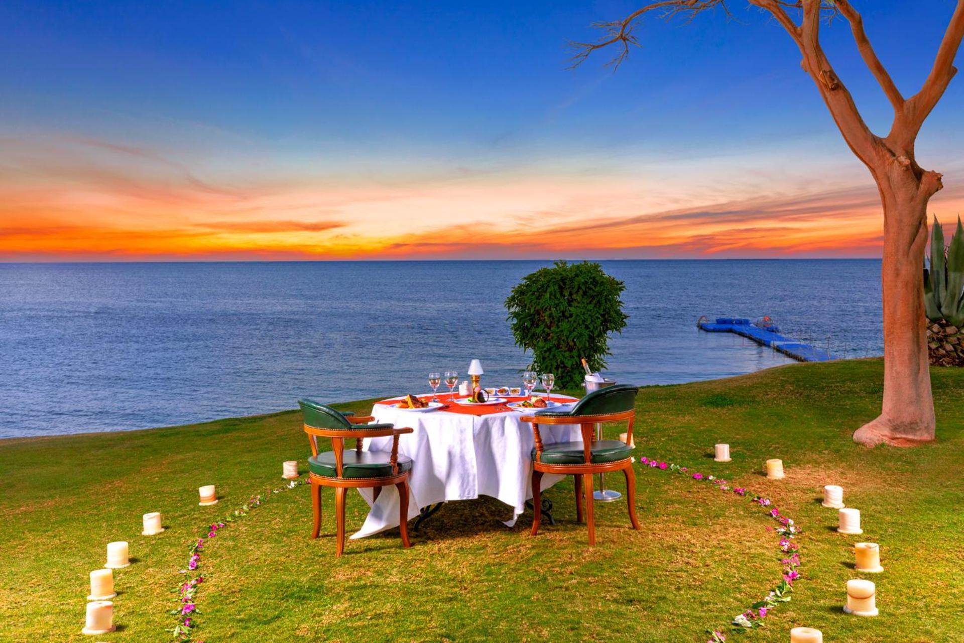 Pickalbatros Royal Grand Sharm - Adults Friendly 16 Years Plus Exteriör bild The photo depicts a romantic outdoor dining setup by the seaside. In the foreground, there is a round table elegantly set with a white tablecloth, plates, and glassware, creating a cozy atmosphere. Surrounding the table are green chairs. A circle of 