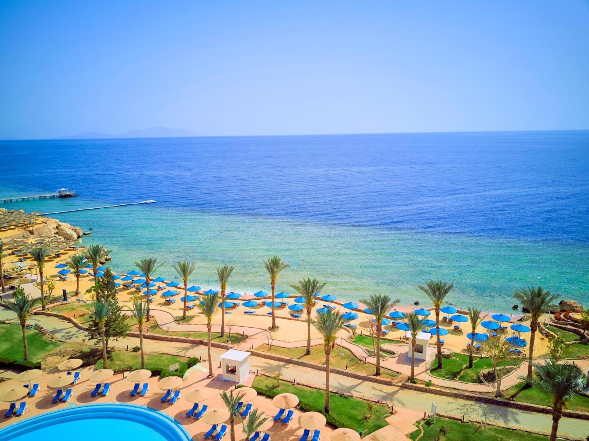 Pickalbatros Royal Grand Sharm - Adults Friendly 16 Years Plus Exteriör bild The photo displays a beautiful coastal scene featuring a clear blue sea that transitions into green tones near the shore. In the foreground, there are sandy beaches lined with palm trees and a variety of beach umbrellas. There is also a swimming pool