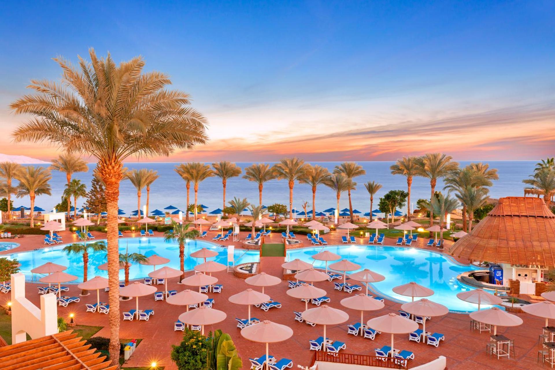 Pickalbatros Royal Grand Sharm - Adults Friendly 16 Years Plus Exteriör bild The image shows a beautiful resort setting by the beach. There is a large swimming pool at the center, surrounded by lounge chairs and umbrellas. Palm trees line the area, giving it a tropical vibe. In the background, you can see the ocean and a colo