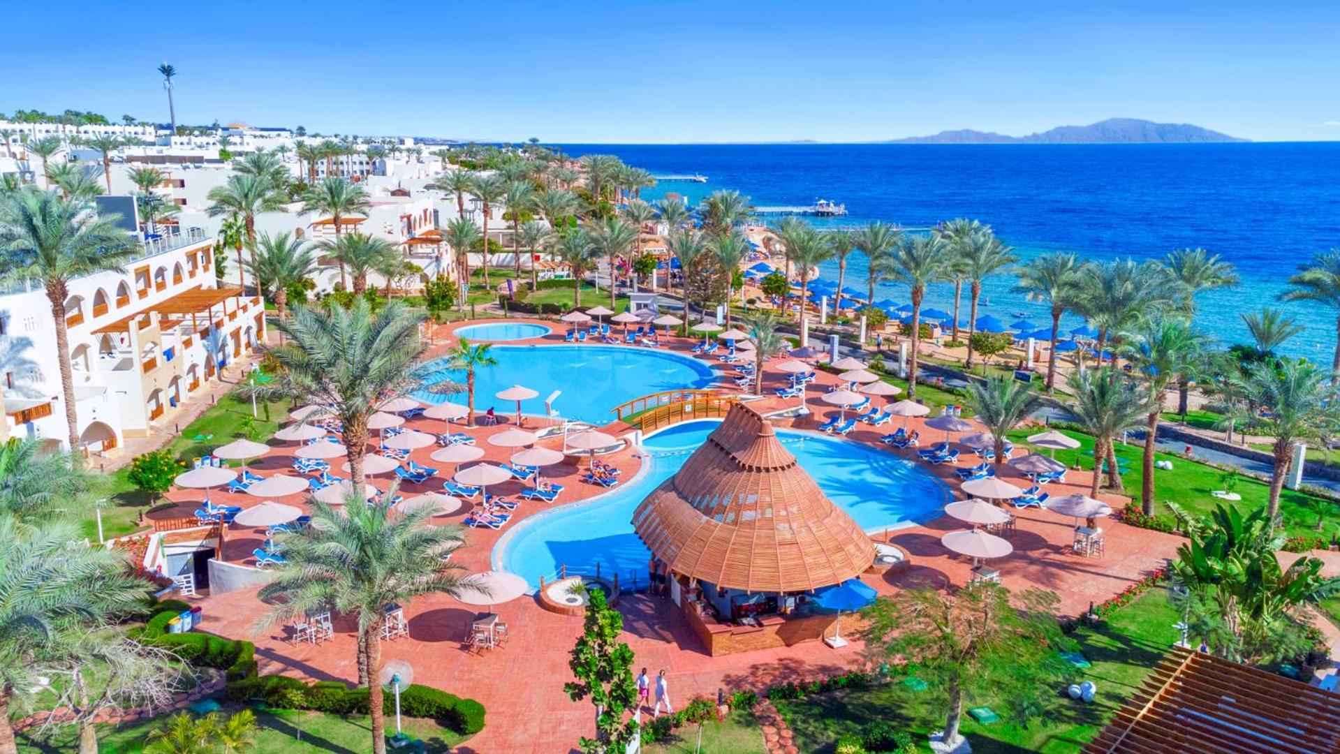 Pickalbatros Royal Grand Sharm - Adults Friendly 16 Years Plus Exteriör bild The image depicts a vibrant resort scene overlooking a clear blue sea. In the foreground, there are multiple swimming pools surrounded by sun loungers and umbrellas. A thatched-roof gazebo is situated near the center of the pool area. Palms and lush 