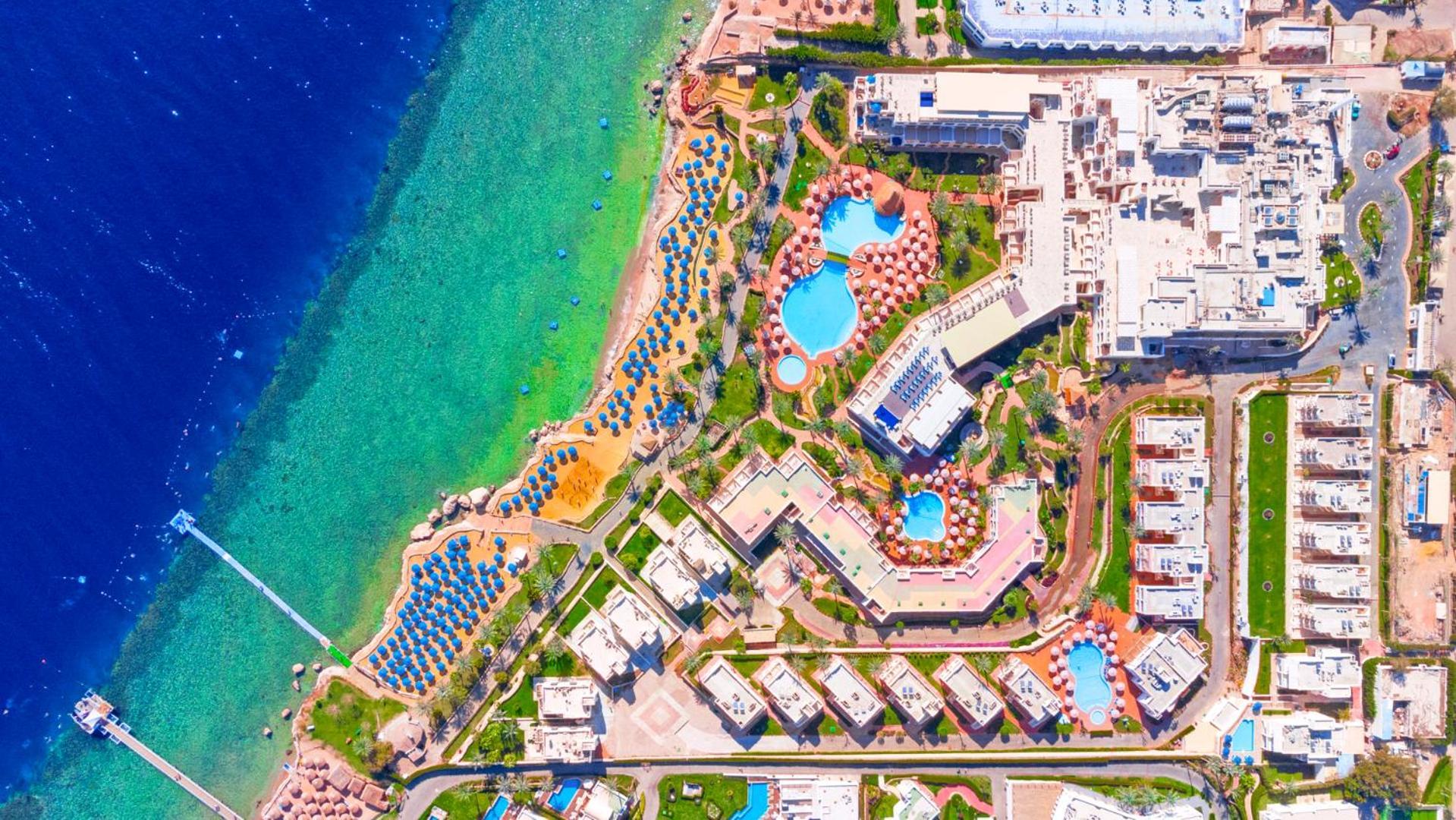 Pickalbatros Royal Grand Sharm - Adults Friendly 16 Years Plus Exteriör bild The image shows an aerial view of a coastal resort area. It features various swimming pools outlined by landscaped gardens and pathways. There are also rows of sun loungers and umbrellas near the beach. The water transitions from a deep blue to a vib