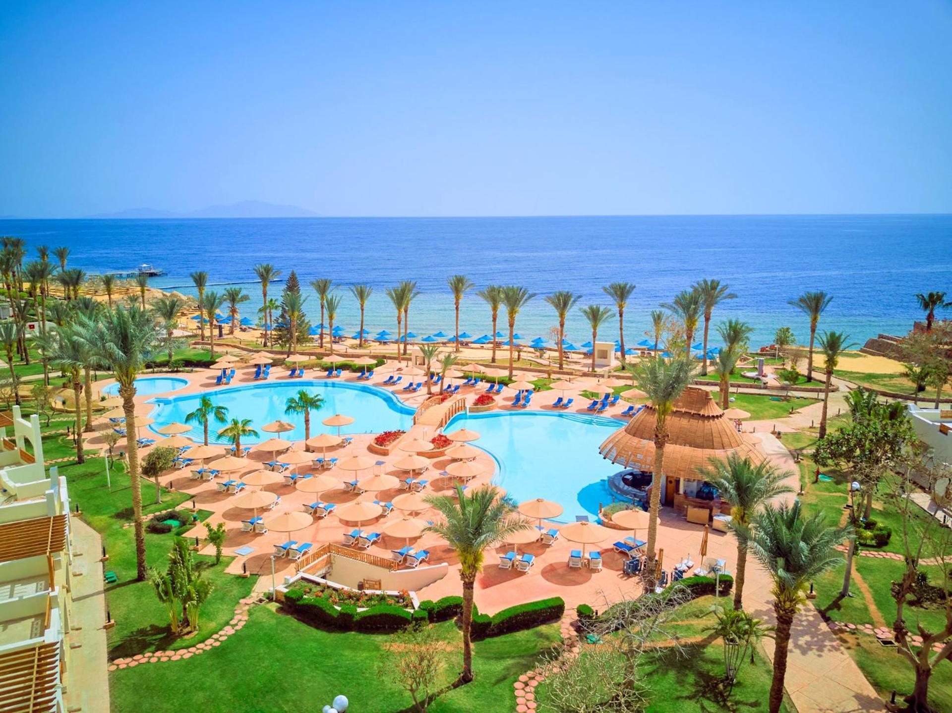 Pickalbatros Royal Grand Sharm - Adults Friendly 16 Years Plus Exteriör bild The photo depicts a beautiful resort scene by the ocean. In the foreground, there is a large swimming pool surrounded by palm trees and lounge chairs. The pool area features sunbeds and umbrellas, suggesting a relaxing atmosphere for guests. In the b