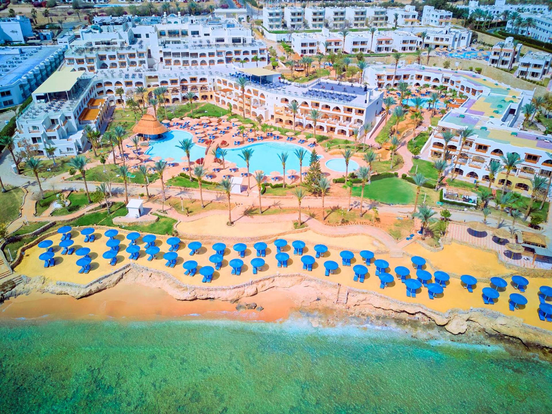 Pickalbatros Royal Grand Sharm - Adults Friendly 16 Years Plus Exteriör bild The photo shows a large resort complex situated near a beach. There is a turquoise swimming pool at the center, surrounded by green lawns and palm trees. Sun loungers with blue umbrellas are lined up on the sandy beach, and the beach area is adjacent