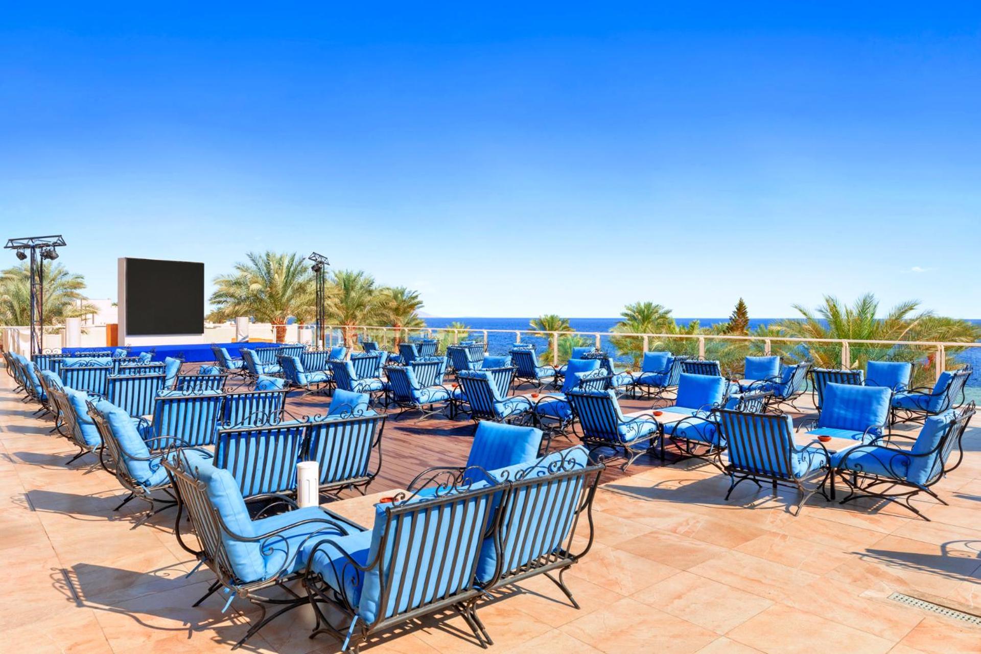 Pickalbatros Royal Grand Sharm - Adults Friendly 16 Years Plus Exteriör bild The photo features a sunny outdoor space with numerous blue lounge chairs arranged in rows on a tiled patio. In the background, there is a clear blue sky and a view of the ocean, along with some palm trees adding to the tropical atmosphere. Additiona